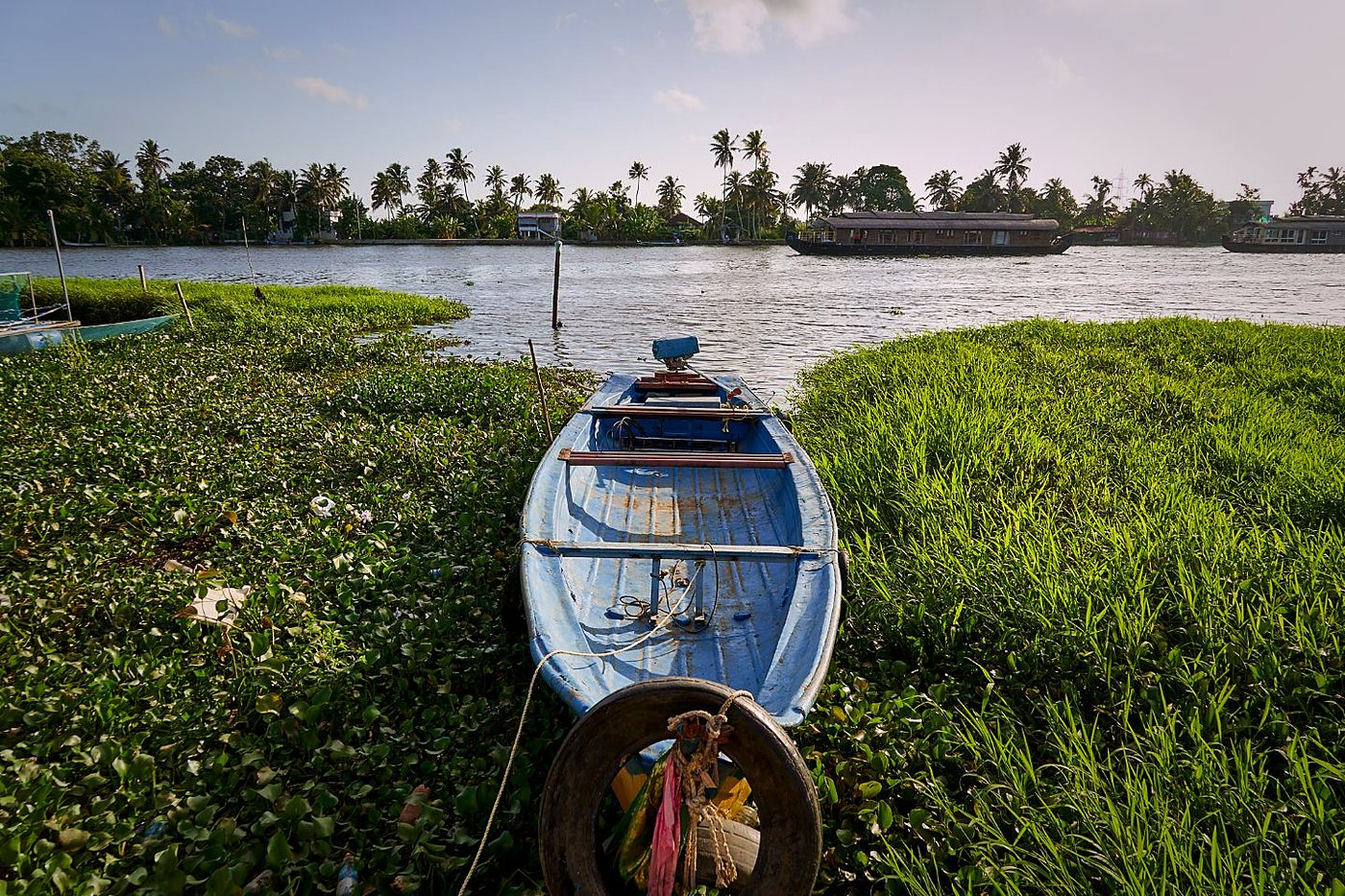 A small boat moored at the edge of the backwaters, houseboats passing in the distance